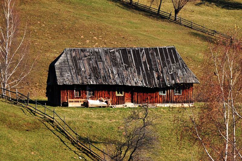 A weathered wooden barn with a dark roof inside a fenced hillside meadow